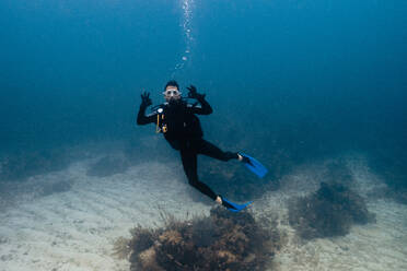 Ein Taucher gibt das OK-Zeichen beim Schwimmen in der Nähe des Meeresbodens, umgeben von blauem Wasser und Meereslebewesen in Cancun, Mexiko - ADSF50774
