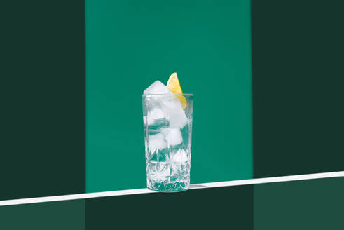 Eine minimalistische Szene mit einer transparenten Tasse, gefüllt mit Gin Tonic und einer Limettenscheibe, vor einem zweifarbigen grünen Hintergrund. - ADSF50780