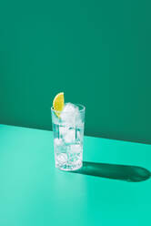 A glass filled with gin tonic and topped with a lime slice stands against a vivid teal backdrop, casting a crisp shadow. - ADSF50782