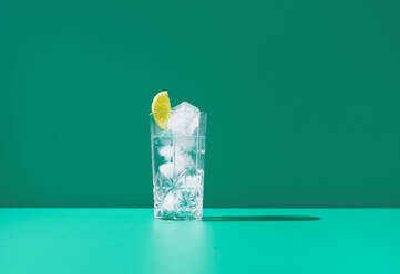 A clear glass full of gin tonic and topped with a lime slice sits against a vibrant green backdrop, creating a fresh and cool visual. - ADSF50783