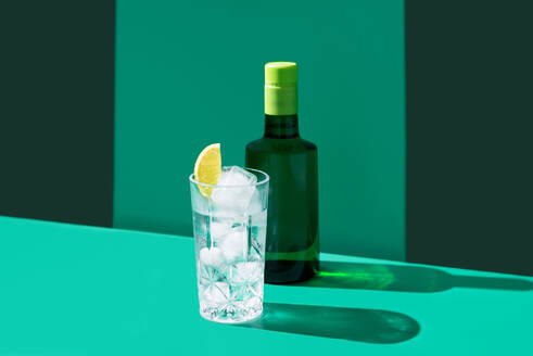 Ein erfrischender Gin Tonic mit Eiswürfeln und einer Limettenscheibe, daneben eine Flasche Gin, vor einem grünen Hintergrund mit Schatten. - ADSF50784