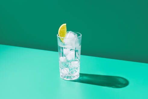 Eine minimalistische Aufnahme eines klaren Glases, gefüllt mit Gin Tonic und einer Limettenscheibe, die einen Schatten auf einen grünen Hintergrund wirft. - ADSF50785