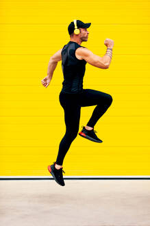 Side view full body of energetic male sportsman wearing headphones and black activewear jumping in air over street against yellow wall - ADSF50786