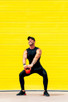 Selbstbewusster, kräftiger männlicher Athlet, der mit einer schweren Kettlebell trainiert und wegschaut, während er auf der Straße über einer gelben Wand steht - ADSF50789