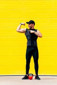 Ganzkörperansicht eines selbstbewussten, lächelnden männlichen Sportlers in Sportkleidung, der seinen Bizeps zeigt und auf ihn zeigt, während er auf der Straße über einer gelben Wand steht - ADSF50790