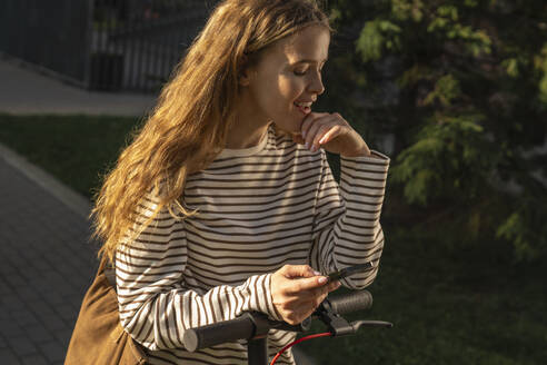 Blonde Frau mit elektrischem Tretroller, die eine Textnachricht auf einem Mobiltelefon schreibt - VPIF09090