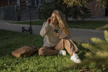 Blonde Frau, die über ihr Smartphone spricht und im Gras sitzt - VPIF09101