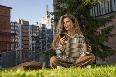 Glückliche Frau mit Mobiltelefon im Gras sitzend im Park - VPIF09102