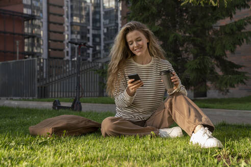 Woman using smart phone sitting on grass in park - VPIF09103