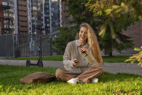 Happy woman with smart phone and disposable cup in park - VPIF09105