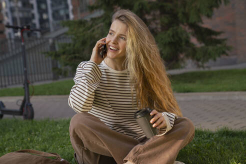 Blonde Frau mit Einwegbecher spricht mit Smartphone im Park - VPIF09106
