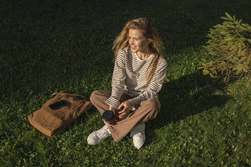 Frau mit Einwegbecher sitzt auf Gras im Park an einem sonnigen Tag - VPIF09107