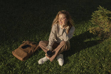 Smiling woman with disposable cup sitting in park at sunny day - VPIF09108