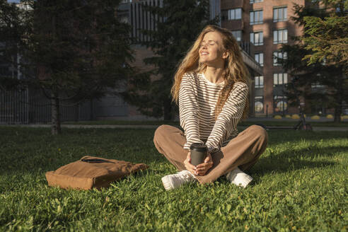 Woman with eyes closed and disposable cup sitting on grass in park - VPIF09110