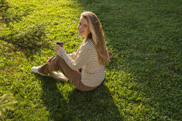 Lächelnde blonde Frau mit Einwegbecher im Gras sitzend - VPIF09111