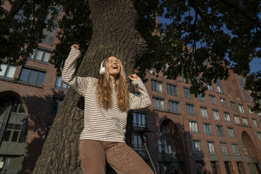Fröhliche Frau, die vor einem Baum steht und über Kopfhörer Musik hört - VPIF09134