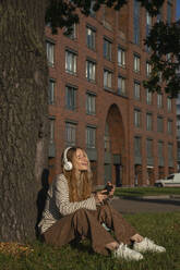 Frau hört Musik über drahtlose Kopfhörer vor einem Baum - VPIF09138