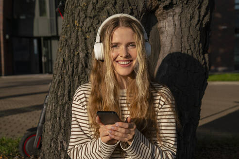 Lächelnde blonde Frau mit Smartphone, die im Sommer über Kopfhörer Musik hört - VPIF09141
