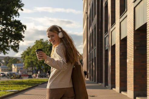 Happy woman with disposable cup listening to music on footpath - VPIF09153