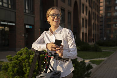 Smiling businesswoman with mobile phone and electric push scooter on footpath - VPIF09172