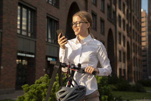 Geschäftsfrau, die ihr Smartphone im Stehen mit einem Elektroroller benutzt - VPIF09174