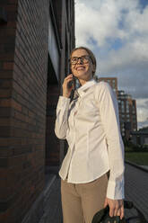 Smiling businesswoman talking over smart phone standing on footpath - VPIF09180