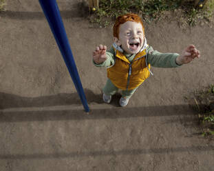 Glücklicher rothaariger Junge mit erhobenen Armen auf dem Spielplatz - MBLF00209