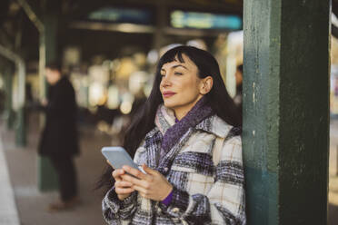 Nachdenkliche Frau mit Smartphone auf dem Bahnsteig stehend - JCCMF10999