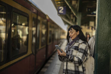 Frau, die ein Smartphone benutzt und am Bahnsteig vor einem ankommenden Zug steht - JCCMF11000