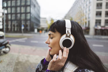 Woman listening to music through wireless headphones on footpath - JCCMF11008