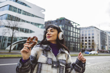 Woman with pretzel listening to music through wireless headphones in city - JCCMF11011