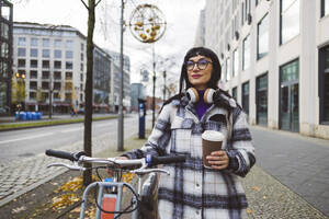 Frau mit Einweg-Kaffeebecher und Elektrofahrrad auf dem Fußweg stehend - JCCMF11013