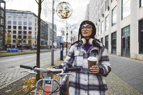 Woman with disposable coffee cup and electric bicycle standing on footpath - JCCMF11013