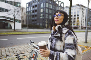 Thoughtful woman with disposable cup and electric bicycle standing on footpath - JCCMF11014