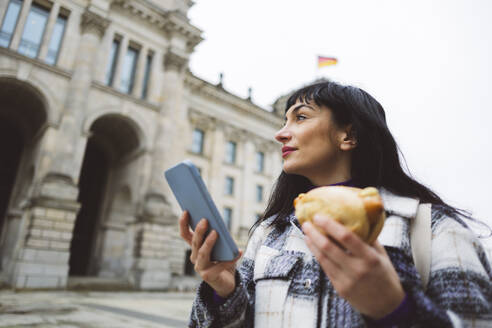 Nachdenkliche Frau mit Smartphone und Brötchen am Gebäude - JCCMF11034