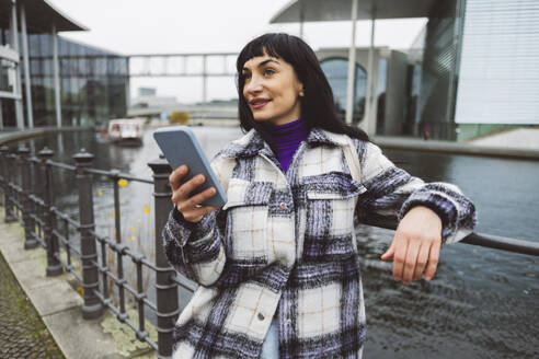 Nachdenkliche Frau mit Mobiltelefon vor einem Fluss stehend - JCCMF11039