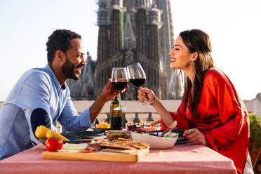 Multirassisches glückliches Liebespaar auf einem Dachbalkon an der Sagrada Familia, Barcelona - Multiethnische Menschen beim romantischen Aperitif-Dinner auf einer Terrasse mit Blick auf die Stadt, Konzepte über Tourismus und Lebensstil der Menschen - DMDF08310