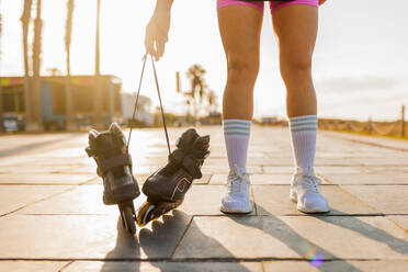 Anonyme Frau mit weißen Turnschuhen und Socken, die schwarze Inline-Skates hält, während sie auf einem Fußweg in einem Park steht, mit Gegenlicht - ADSF50792