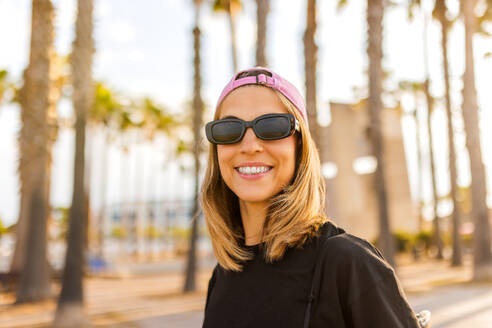 Porträt einer lächelnden attraktiven jungen Frau mit schwarzer Sonnenbrille und T-Shirt mit rosa Kappe vor unscharfem Hintergrund im Park am Wochenende - ADSF50793