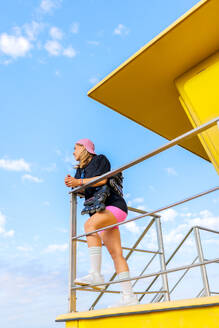 Ganzkörperaufnahme einer jungen kaukasischen Frau in Freizeitkleidung mit Inline-Skates, die sich am Geländer einer Rettungsschwimmerhütte am Strand unter blauem Himmel anlehnt - ADSF50794