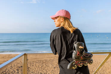 Back view of young Caucasian woman dressed in casuals with inline roller skates leaning on lifeguard hut railing at beach under blue sky - ADSF50797