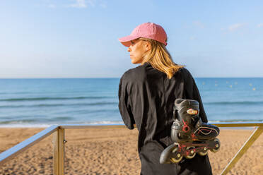 Rückenansicht einer jungen kaukasischen Frau in Freizeitkleidung mit Inline-Skates, die sich am Geländer einer Rettungsschwimmerhütte am Strand unter blauem Himmel anlehnt - ADSF50797