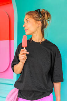Portrait of beautiful Caucasian young woman with pink ice cream popsicle while standing against blue wall looking away - ADSF50807
