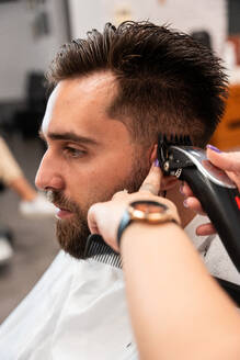 Abgeschnittene, nicht erkennbare Friseurin bei der Verwendung eines elektrischen Trimmers an den Haaren eines männlichen Kunden im Salon - ADSF50813