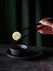 Sushi roll held by anonymous person using chopsticks with a droplet of sauce ready to fall in bowl placed on table against green curtains - ADSF50814
