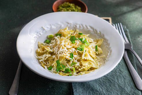 A white bowl filled with Farfalle pasta, peas, and grated cheese, accompanied by a pesto sauce container, placed on a dark surface with cutlery alongside. - ADSF50826