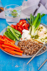 Eine lebendige und nahrhafte Frühstücksplatte mit Kirschtomaten, Gurken, grünen Zwiebeln, rotem Paprika, Fetakäse, Buchweizen und Spiegeleiern, die mit Pesto beträufelt sind. - ADSF50829