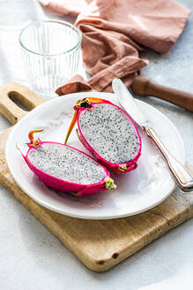 Eine leuchtend rosafarbene Drachenfrucht, die in der Mitte durchgeschnitten ist und ihr gesprenkeltes Inneres preisgibt, wird auf einem weißen Teller mit einem rustikalen Holzbrett präsentiert. - ADSF50830