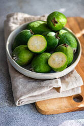 Rohe Feijoa-Früchte, die auch als Ananas-Guaven bekannt sind, werden in einer Keramikschale auf einem Holzbrett mit Leinentuch präsentiert. - ADSF50831