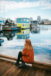 Eine nachdenkliche rothaarige Frau sitzt auf einem hölzernen Steg und blickt auf das ruhige Wasser, das von lebhaften schwimmenden Häusern in Victoria, Vancouver Island, British Columbia, umgeben ist. - ADSF50837